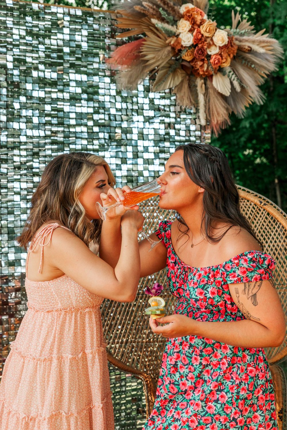 Two friends drinking cocktails against metallic backdrop with dried floral arrangement.