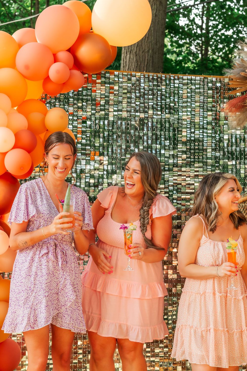 Friends laughing together at outdoor party with orange balloon arch and sparkly backdrop.