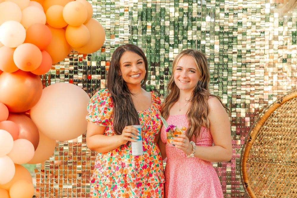 Party scene with orange balloon arch and sparkly backdrop as friends celebrate with drinks.