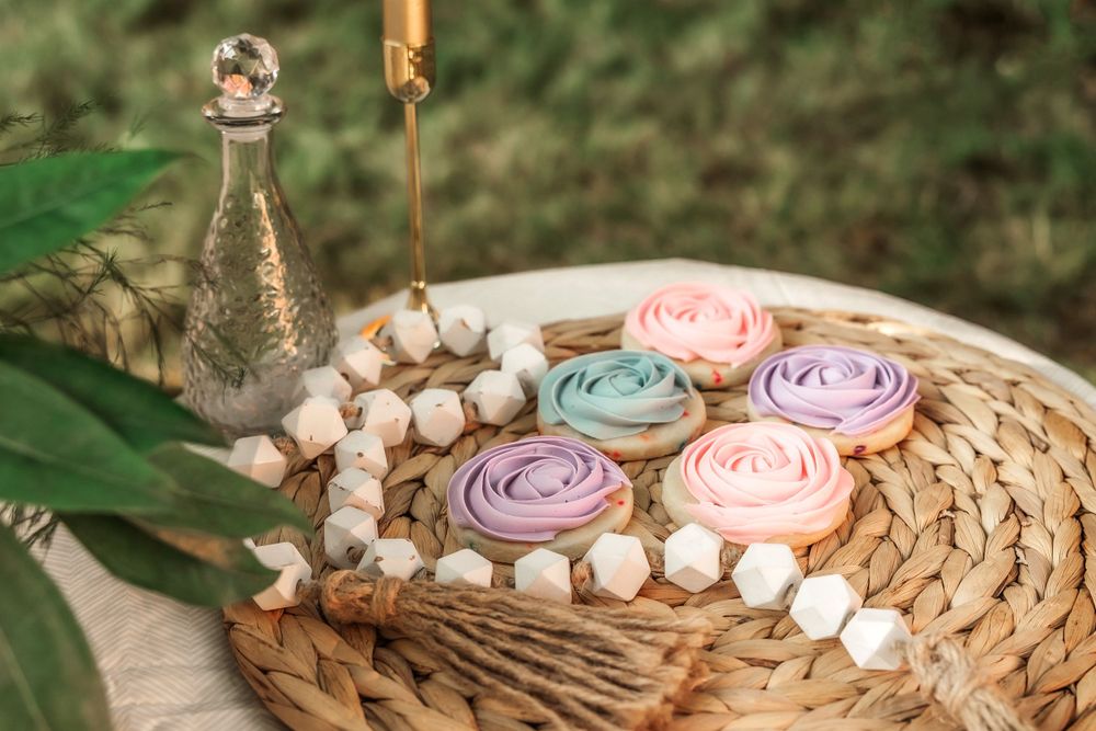 Pastel colored cupcakes with delicate frosting roses displayed on a woven platter with cotton stems and decorative bottle.