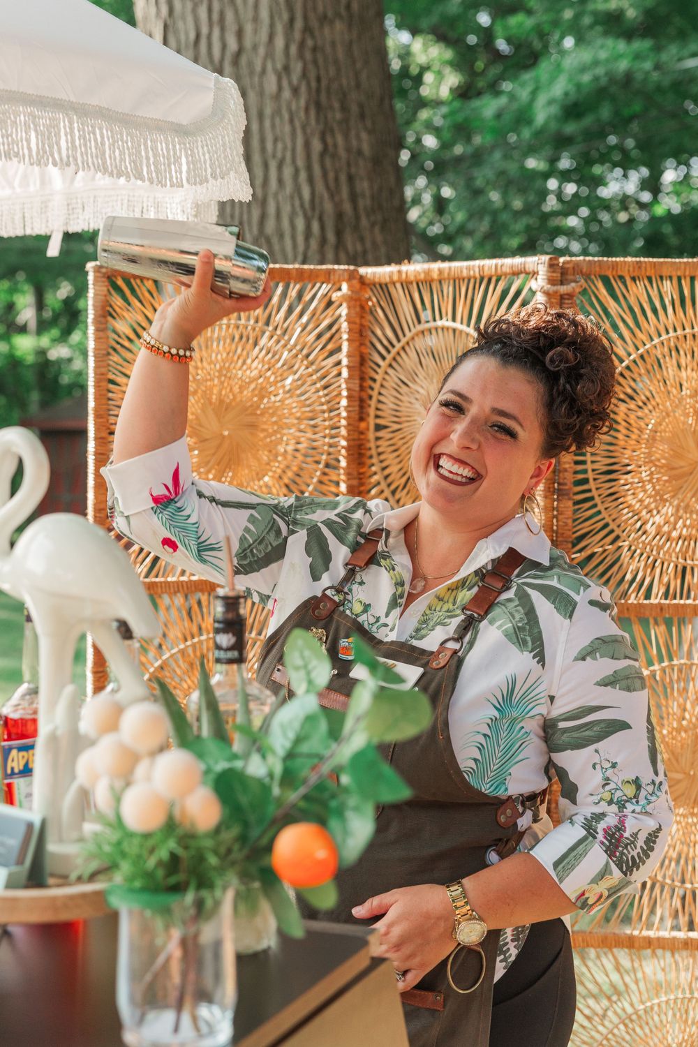 Bartender in tropical print shirt mixing drinks at outdoor event with rattan decor.
