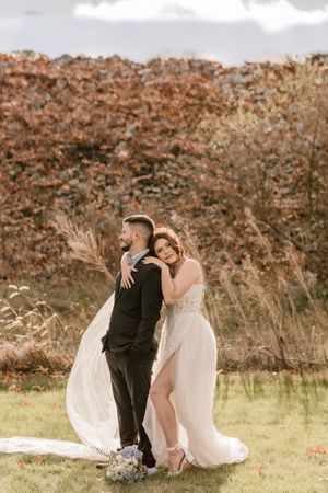 A romantic wedding couple sits together in a sunlit field with autumn foliage in the background.