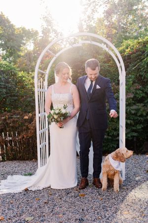 A couple poses under a white garden arch with their dog at their outdoor wedding ceremony.