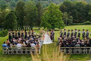 Panoramic view of an outdoor wedding ceremony with guests seated on white chairs surrounded by lush green landscape.