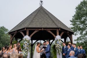 Wedding ceremony takes place under a wooden gazebo decorated with floral arrangements.