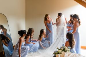 Bridesmaids in light blue dresses help with wedding preparations in a sunlit room.
