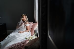 A bride in a white wedding dress sits by a window holding a bouquet, creating a romantic silhouette.