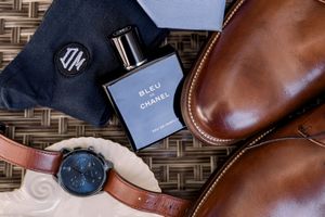 Luxury men's accessories including leather wallet, watch, and sunglasses arranged on textured woven surface.