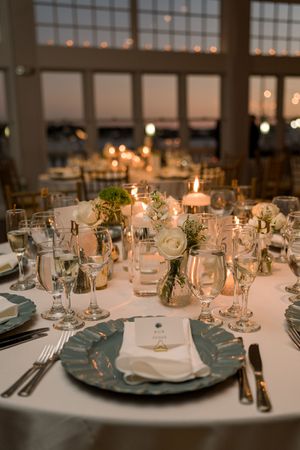 Elegant wedding reception table setting with glowing candles, crystal glasses and blue plates in warm evening light.