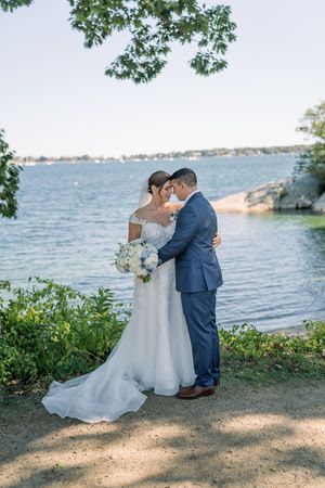 A romantic wedding couple embraces by a scenic waterfront location under tree branches on a sunny day.