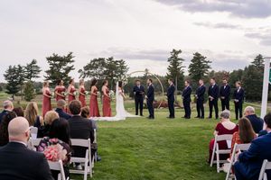 Guests seated in white chairs on a grassy hilltop during an outdoor wedding ceremony with cloudy skies overhead.