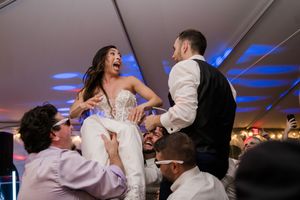 A celebration scene at a wedding reception with guests lifting the couple on chairs under blue lighting.