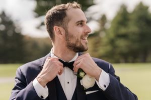 A groom in a navy suit laughs while standing outdoors in a grassy field on a sunny day.
