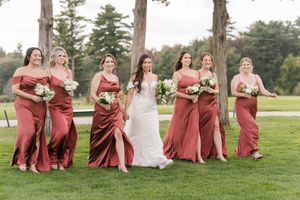 Wedding party wearing elegant rust-colored bridesmaid dresses walk together on a lush green lawn at an outdoor venue.