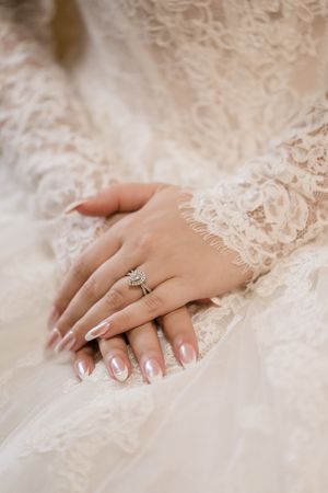Close-up of a bride's manicured hand with engagement ring resting on delicate white lace fabric.