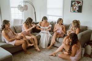 A group in matching blush-colored robes sits together on a gray couch in a bright, sunlit living room.