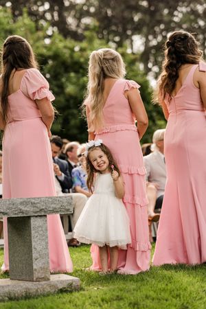 A group of bridesmaids in pink dresses and a flower girl in white sit together on stone blocks in a garden setting.
