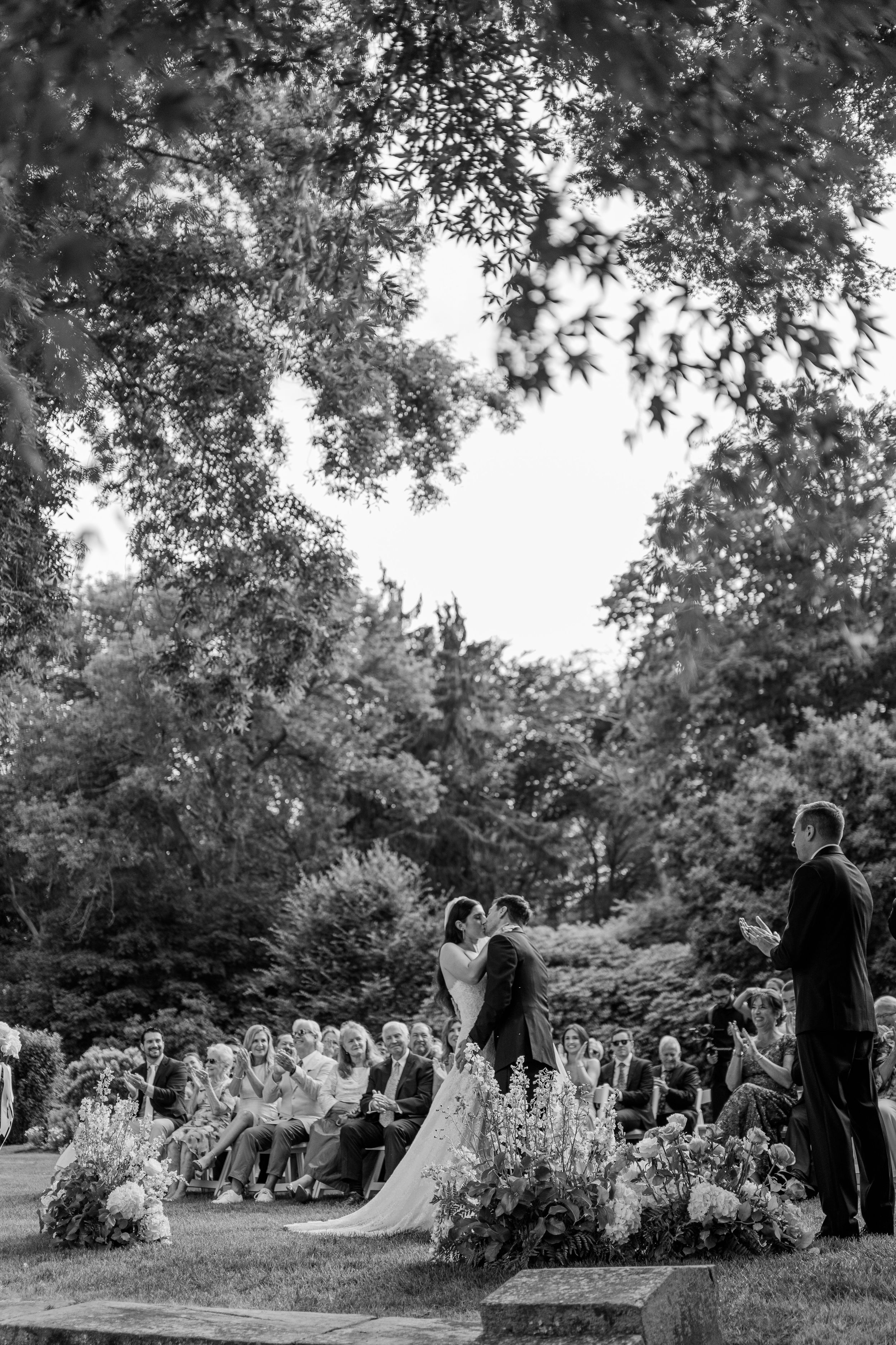 Black and white photo of an outdoor wedding ceremony under trees with guests seated on both sides.