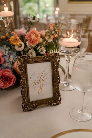 Elegant wedding table setting with ornate gold frame displaying Table 8, coral roses, and glowing candlelight.