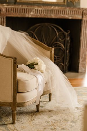 A delicate white wedding veil drapes over an elegant cream-colored French chair beside an ornate fireplace.