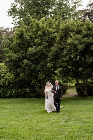 A couple walks together across a lush green lawn with trees in the background.