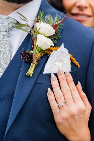 Close-up of white rose boutonniere on navy suit with engagement ring display.