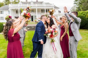 Wedding party celebrates with raised bouquets in front of white colonial-style mansion.