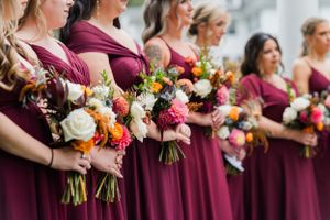 Bridesmaids in burgundy dresses hold vibrant fall-colored bouquets featuring roses and mixed flowers.