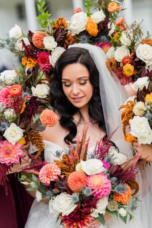 Close-up of a bridal bouquet with coral, burgundy, and white blooms surrounded by autumn foliage.