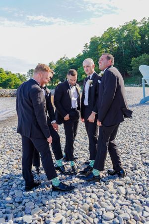 Groomsmen in black tuxedos showing off their matching teal socks on rocky beach.