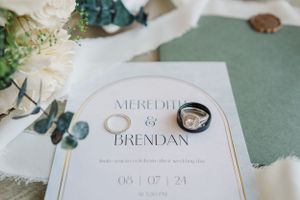 Wedding invitation and rings styled on sage green background with white flowers.
