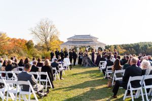 Outdoor wedding ceremony setup with white chairs and fall foliage in the background.