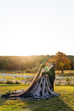 Couple embracing in a scenic autumn field at sunset with their dramatic wedding attire.