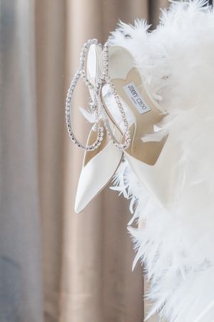 White Jimmy Choo bridal shoes adorned with crystals displayed against a feathered white dress background.