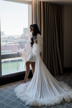 A dramatic silhouette of someone in a flowing white robe with feathered trim looking out a high-rise window at the city.