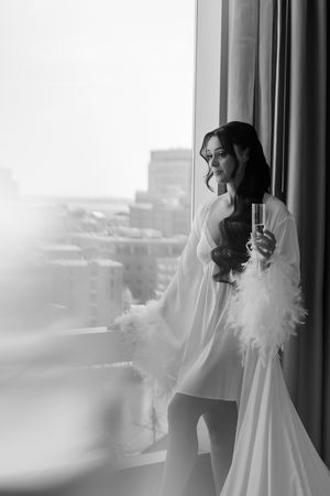 A black and white boudoir photo of a woman in a white robe with feather accents gazing out a high-rise window.