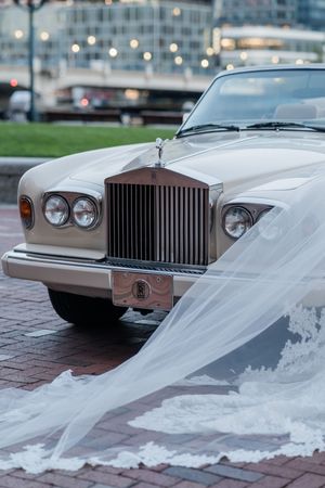 White wedding veil draped over the front of a classic Rolls-Royce luxury car with its iconic grille and headlights.