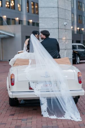 A wedding couple sits in a vintage white car with a flowing lace veil trailing behind them on a brick courtyard.
