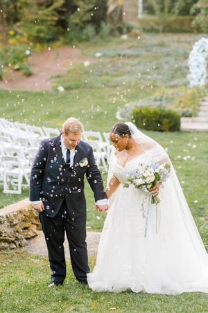 A wedding couple bends down to pet something on the grass while flower petals fall around them during an outdoor ceremony.