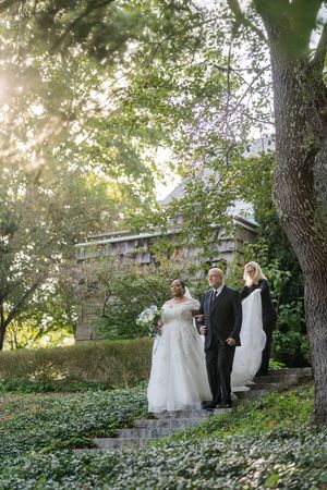 Wedding party gathered on stone steps in a lush garden setting with sunlight filtering through trees.