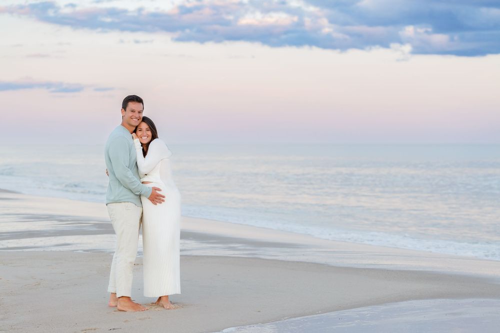 A couple in white attire poses for romantic beach wedding photos at sunset along the ocean shore.