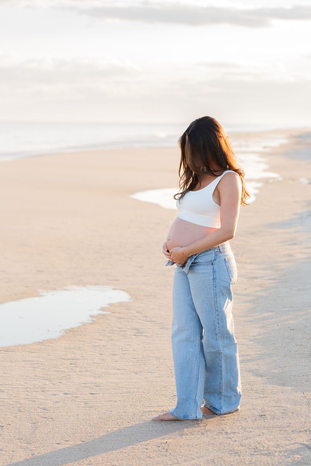 Person in white top and light blue jeans walking along sandy beach at sunset with waves rolling in.
