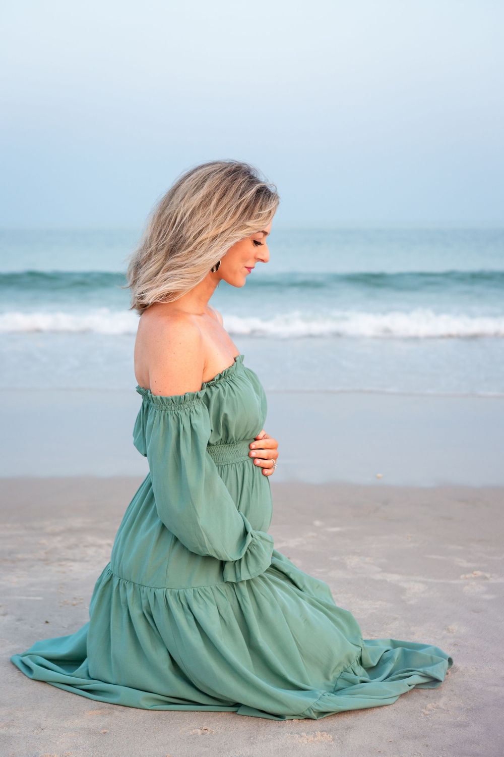 Woman in flowing sage green dress poses gracefully on sandy beach at sunset with ocean waves in background.