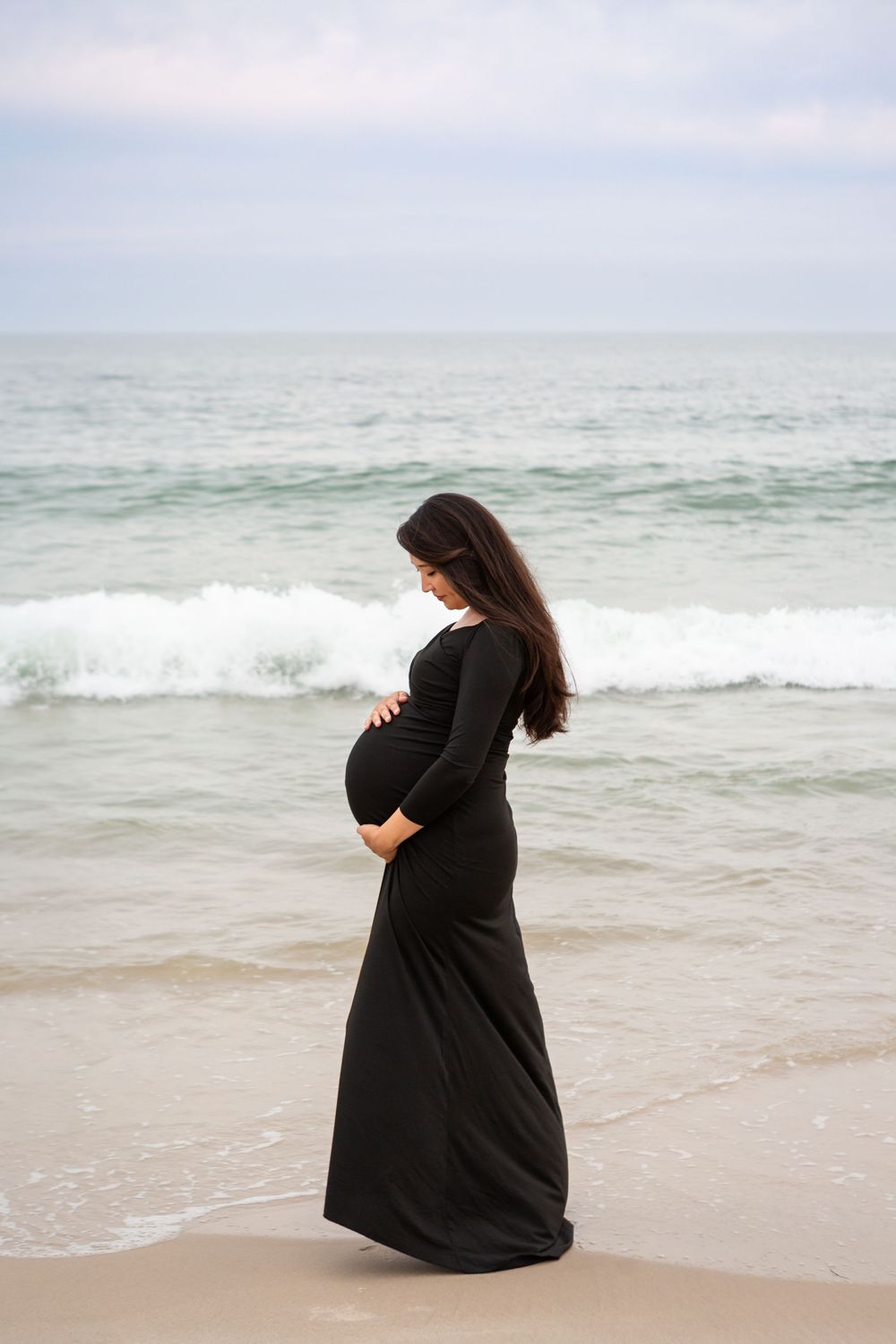 A person in a black dress sits alone on a sandy beach looking out at the ocean waves.