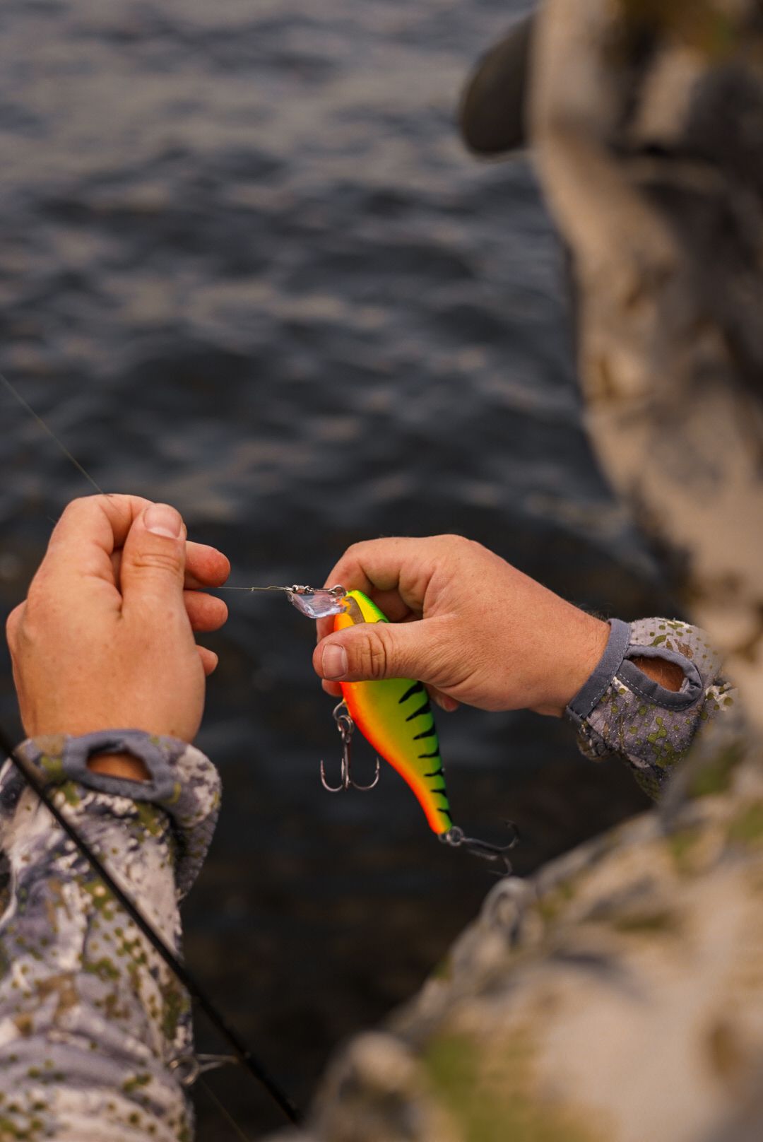 Hands holding a colorful fishing lure against a blurred outdoor background.