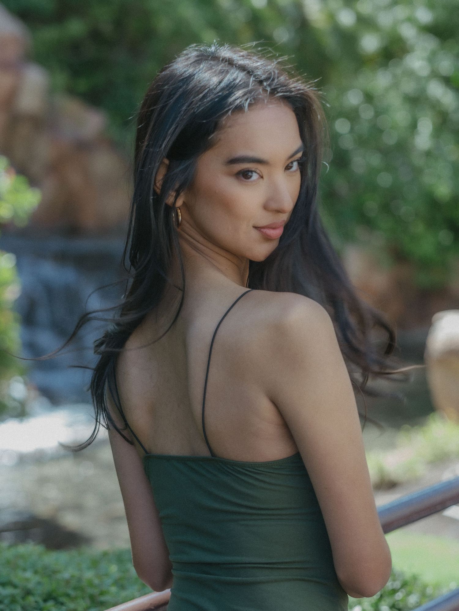 Young woman in a green spaghetti strap dress posing outdoors near a waterfall with lush greenery.