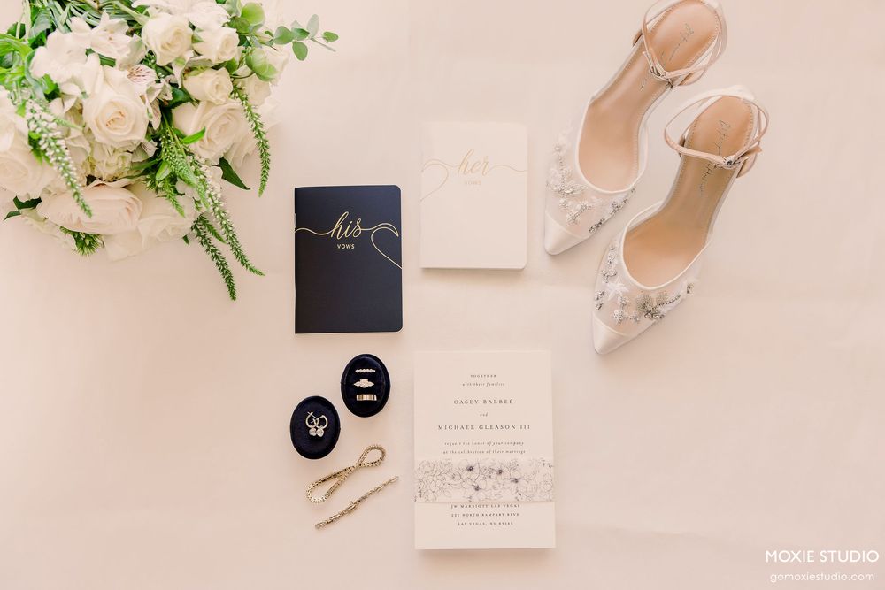 Wedding accessories including white heels, invitation, and floral decorations arranged on light pink background.