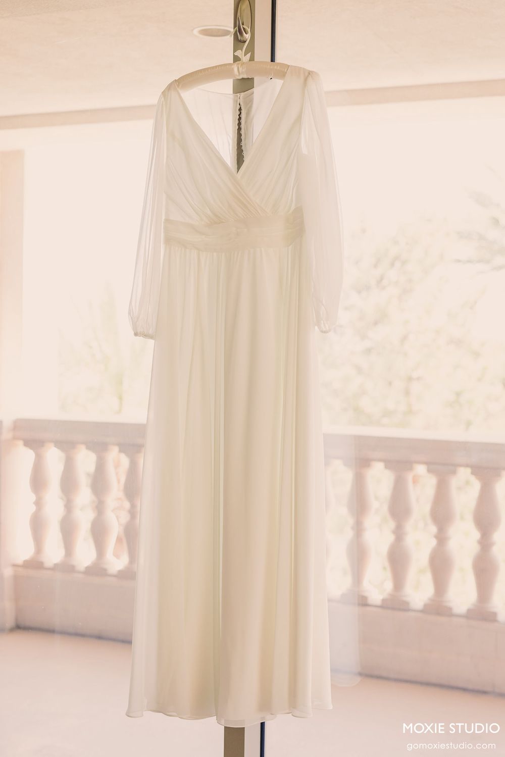 Elegant white chiffon dress displayed on a hanger against a bright window with decorative balustrade railing.
