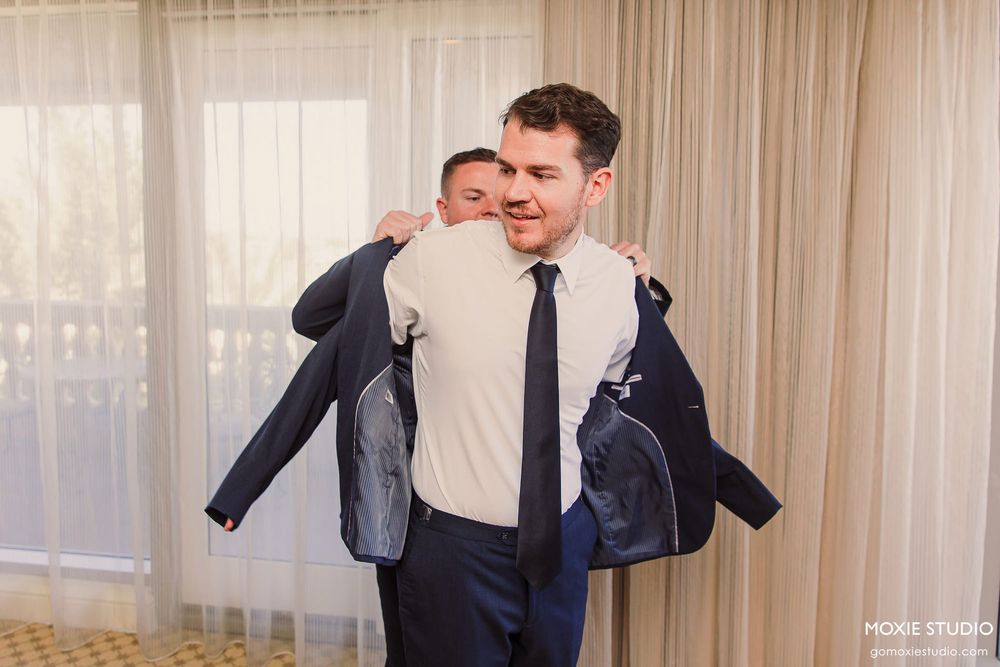 A groom gets ready for his wedding day by putting on a navy suit jacket in a sunlit hotel room.