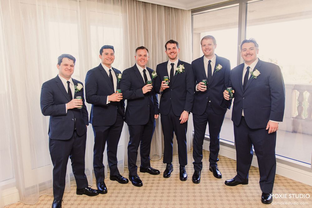 Groomsmen in dark suits pose together in a light-filled room during a wedding celebration.
